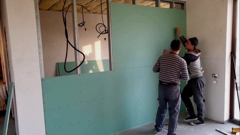 Doi montatori poziționează o placă verde de gips-carton pe structura metalică a unui perete