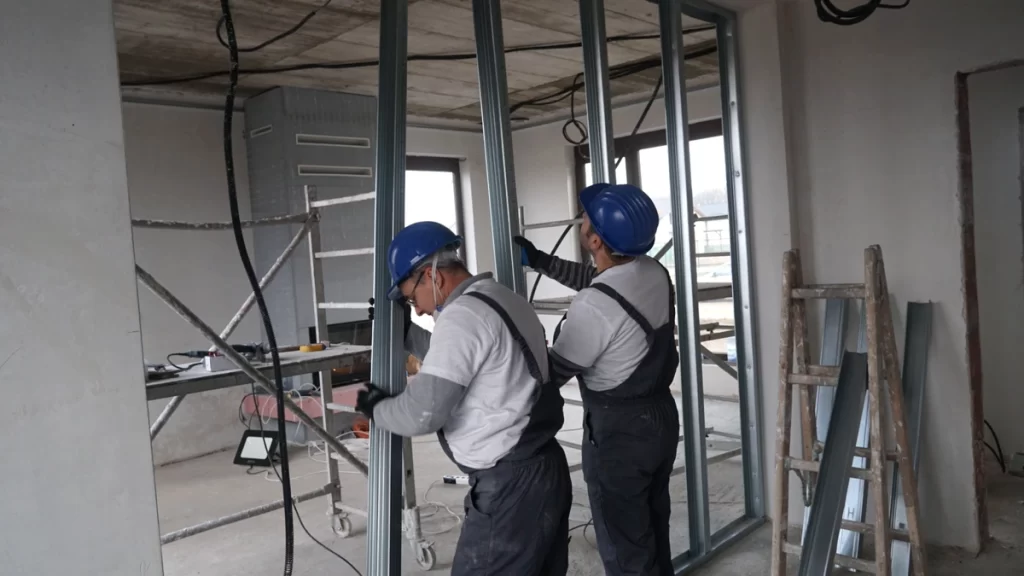 Doi montatori lucrează la structura metalică a unui perete de compartimentare, cu traseele electrice vizibile.