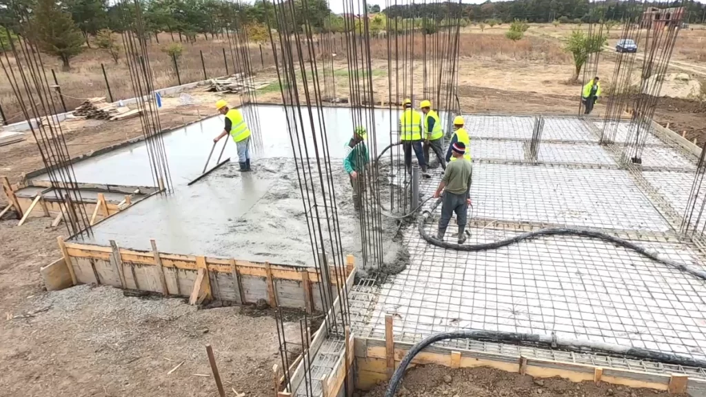 Echipă de muncitori toarnă beton cu pompa și nivelează suprafața unei plăci armate pe șantier.